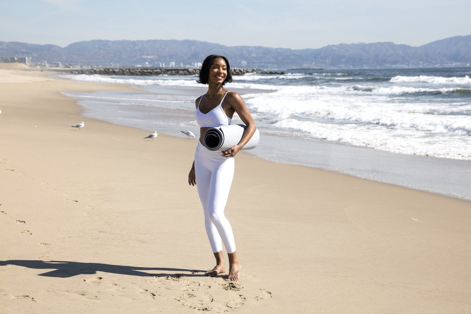 Maya smiling on the beach, holding her yoga mat